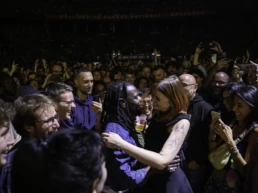 Eric Burton of Black Pumas standing in the crowd at Forest National, Brussels, smiling and embracing a female fan as the audience cheers and captures the moment on their phones.