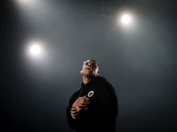 Vince Staples performing at Best Kept Secret Festival in Hilvarenbeek, Netherlands. He looks upward under three soft white spotlights, wearing a black hoodie and glasses, hands clasped near his chest. Photo by Marvin Anthony.