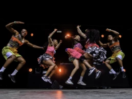 Sho Madjozi performing with her dance crew at Couleur Café Festival in Brussels, Belgium. All six performers are captured mid-jump in colorful outfits against a black stage backdrop with red lights. Photo by Marvin Anthony.