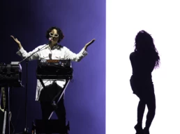 Charlotte Adigéry and Bolis Pupul performing at Lokerse Feesten in Lokeren, Belgium. Bolis stands behind a synthesizer in a white shirt and white sunglasses under blue lighting, while Charlotte’s silhouette appears in profile against a bright white background. Photo by Marvin Anthony.