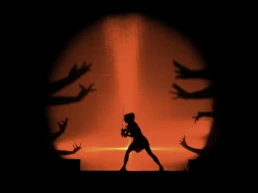 Lindsey Stirling performing at Vorst Nationaal in Brussels, Belgium. She stands in silhouette with a violin, framed by a dramatic red spotlight and theatrical hand shadows stretching toward her from both sides. Photo by Marvin Anthony.